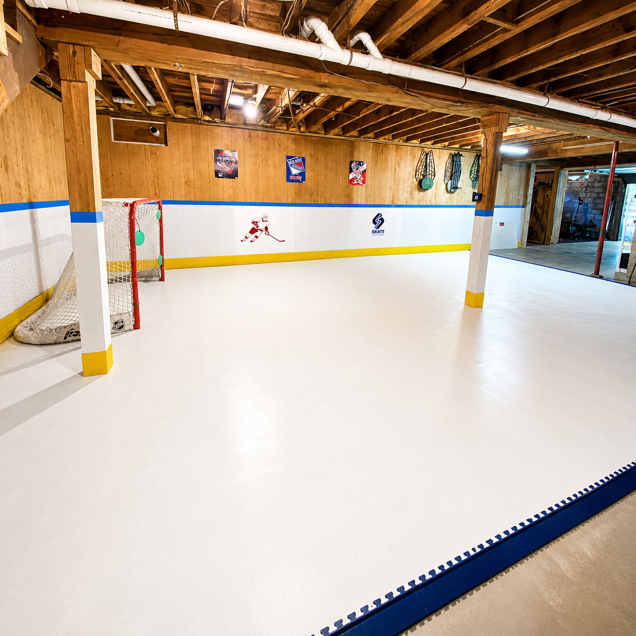 An indoor ice rink or hockey court with a white ice surface, blue and white boards, yellow striping, wooden ceiling beams, and sports wall decals. A hockey goal is visible on the left side.