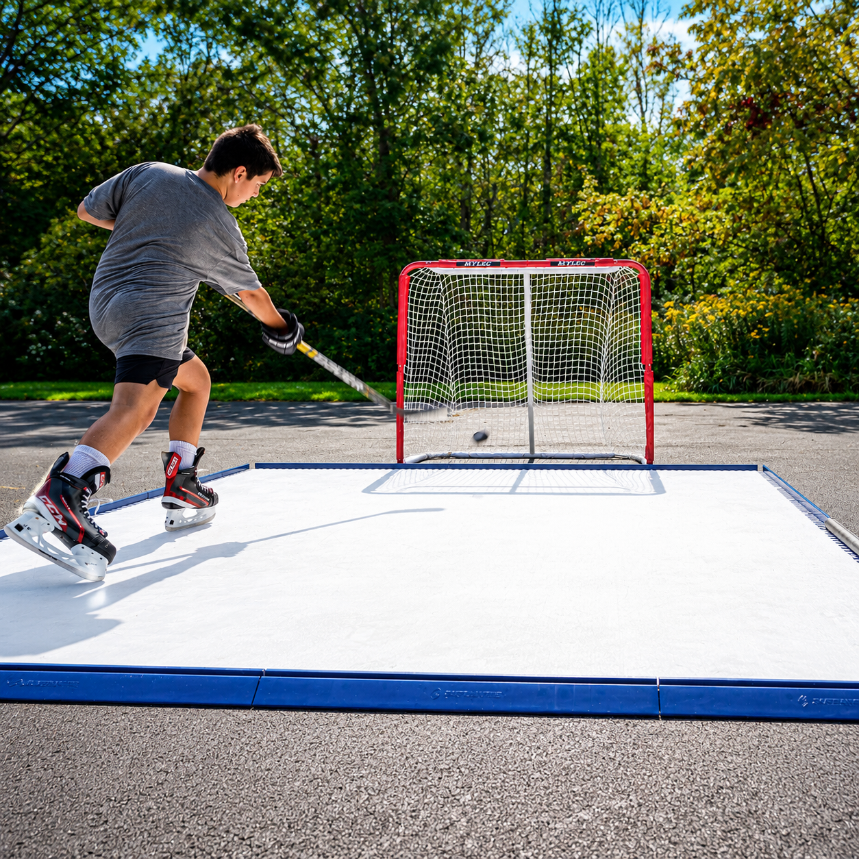 A person ice skating on an outdoor ice rink with a red hockey goal and net, surrounded by trees and grass in a park setting.