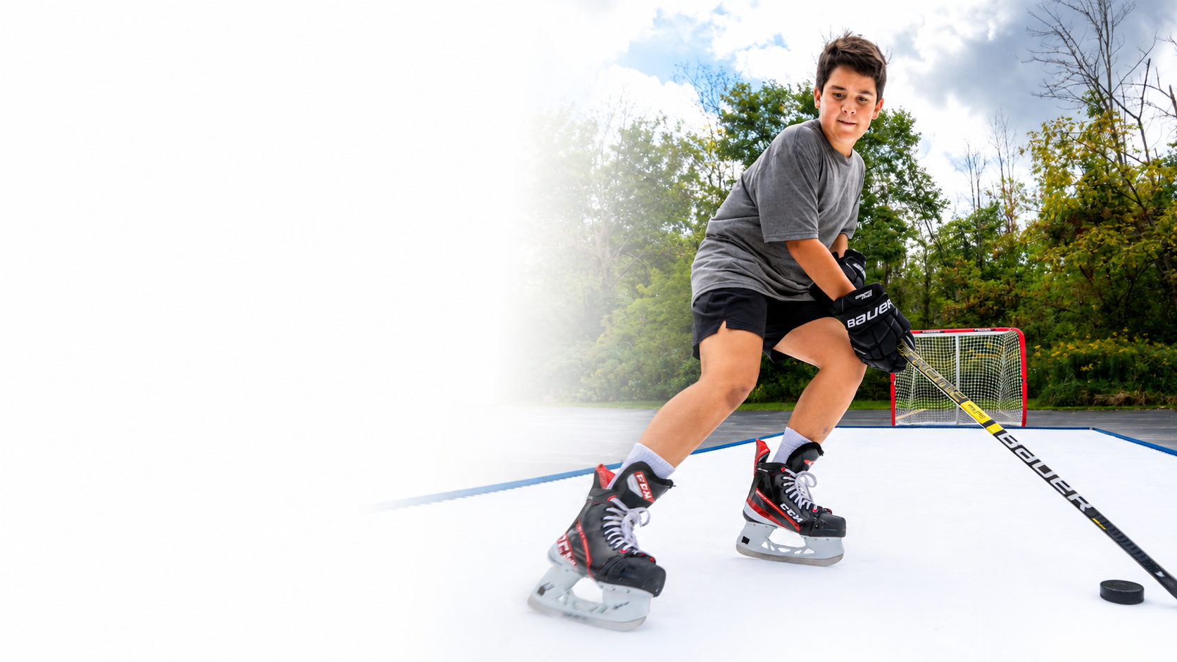A boy wearing a gray t-shirt, black shorts, and hockey gear including Bauer skates and a stick, posed on an outdoor ice rink with trees and a goal in the background.